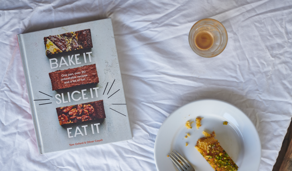 The Exploding Bakery cookbook, BAKE IT, SLICE IT, EAT IT, on a table with some Lemon Polenta & Pistachio cake on a plate.