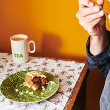 A slice of Hummingbird cake with pecans on top On a green plate with a cup of tea in the background.