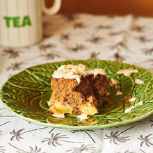 A slice of Hummingbird cake with pecans on top On a green plate with a cup of tea in the background.