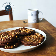 Plate of Hazelnut, Miso & White Chocolate Cookies on a wooden table with a mug in the background, featuring the 'Cake Drops' logo.