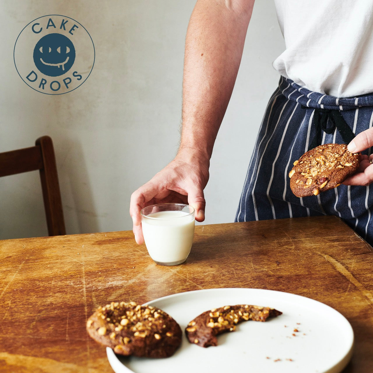 Person holding a Hazelnut, Miso & White Chocolate Cookie and a glass of milk on a wooden table with 'Cake Drops' logo.