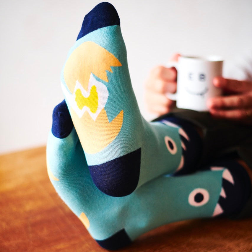 Person wearing Exploding Bakery Socks with a design, sitting at a table with a slice of cake and a mug.