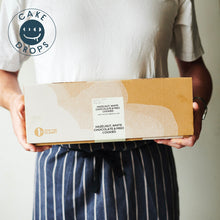 Person holding a box of Hazelnut, Miso & White Chocolate Cookies with 'Cake Drops' branding.