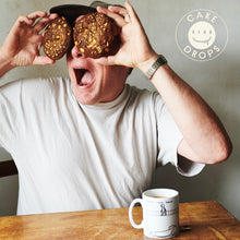 Person holding Hazelnut, Miso & White Chocolate Cookies over their eyes with a mug on a table, featuring the 'Cake Drops' logo.